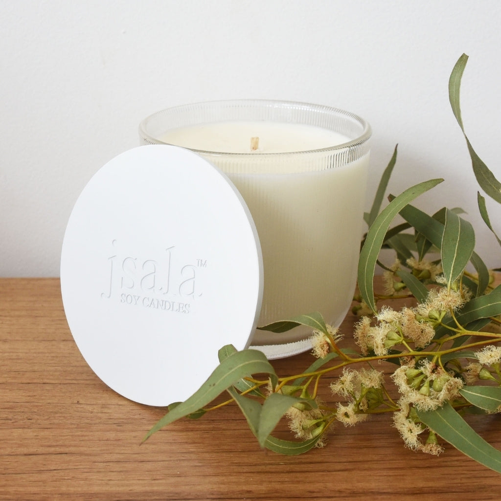 Jsala ribbed glassware candle sitting on table with white lid leaning against it. Sprig of wattle placed next to it.