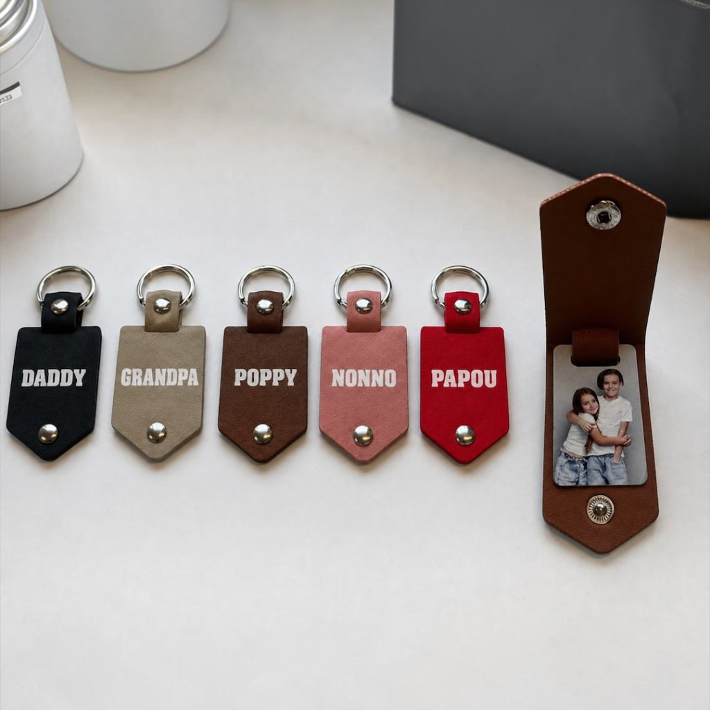 Personalised  leather keychains in black, beige, brown, pink, red and teal. Opens to show two photos inside. The keychains have metal key rings and snap buttons, and the names DADDY, GRANDPA, POPPY, NONNO, and PAPOU are printed on the front.
